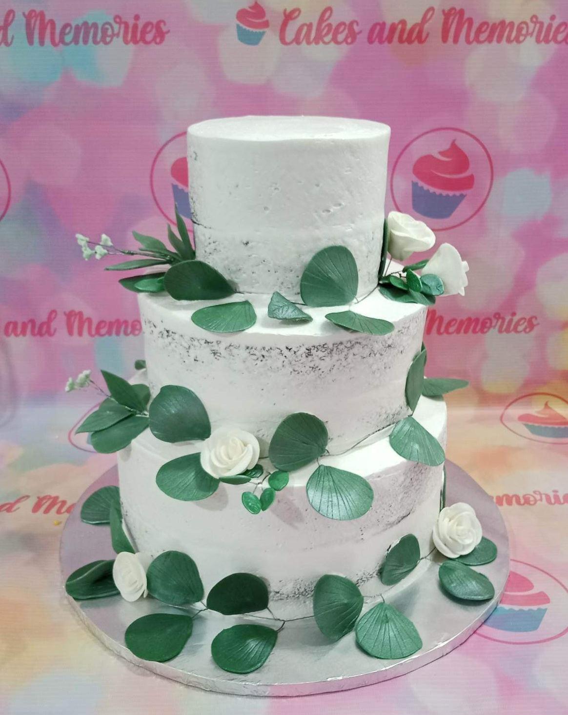 Three-tier semi-naked white wedding cake decorated with green eucalyptus leaves and white gumpaste roses on a silver board.