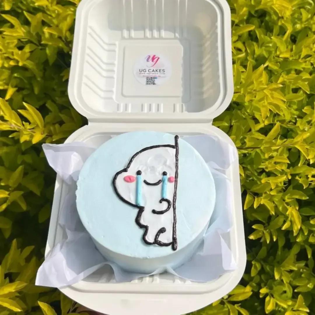 Light blue bento cake with a hand-piped white character peeking from behind a black line, featuring pink cheeks and blue tears.