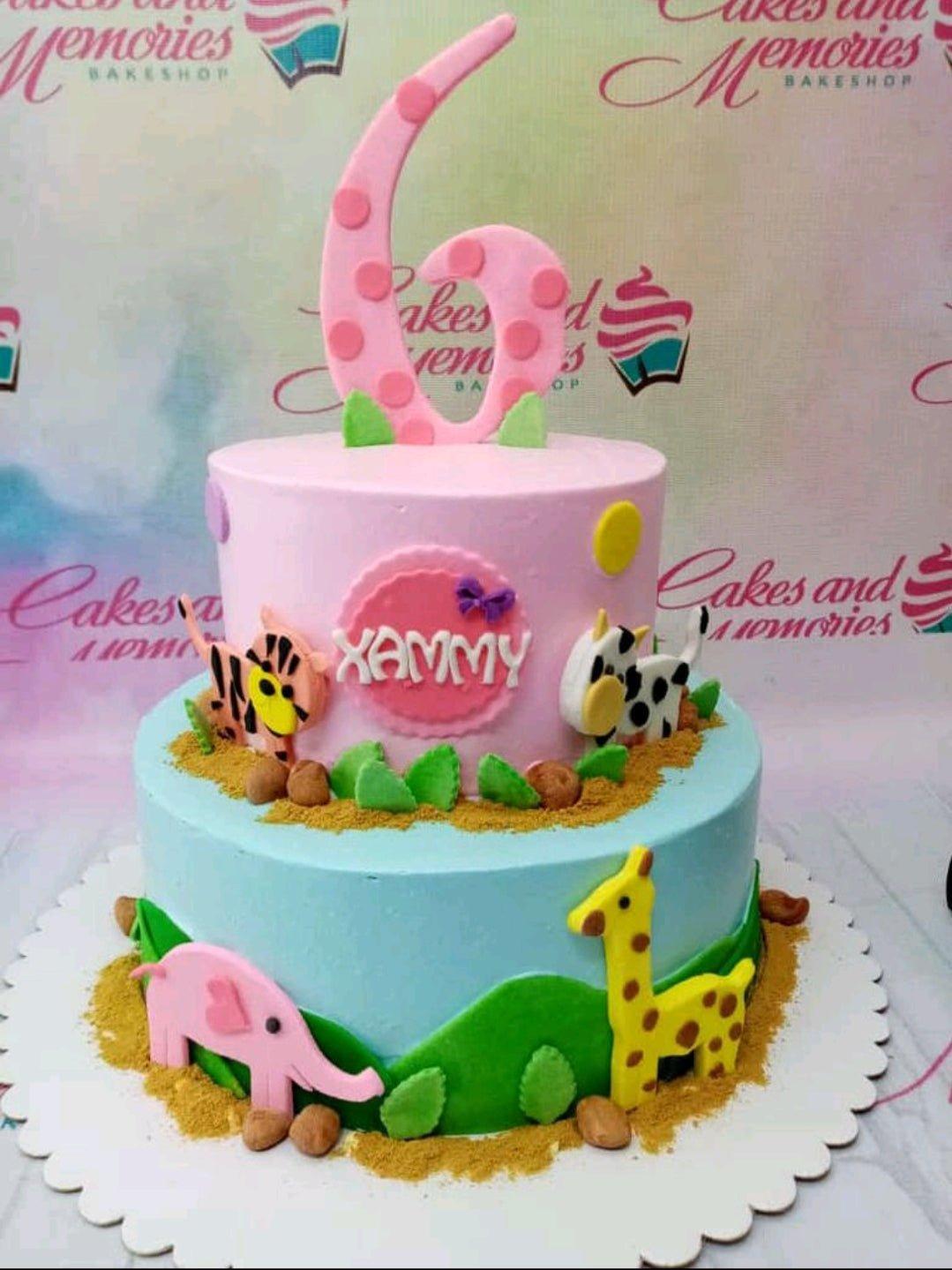 Two-tier safari themed birthday cake with pink and blue icing, featuring fondant animals like a giraffe, elephant, tiger, and cow.