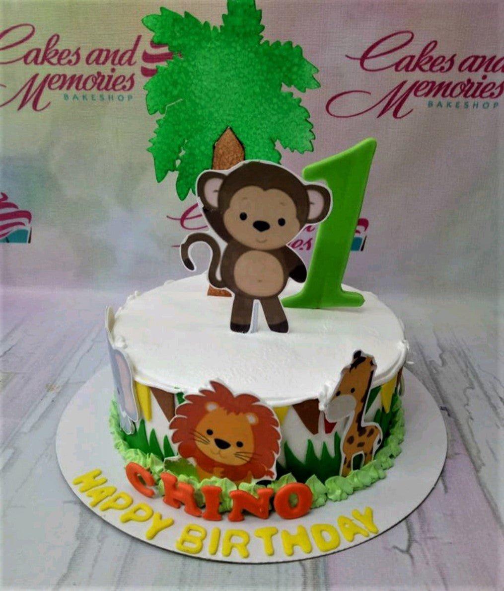 Safari themed 1st birthday cake with monkey, lion, and giraffe printouts on white icing with green grass borders.