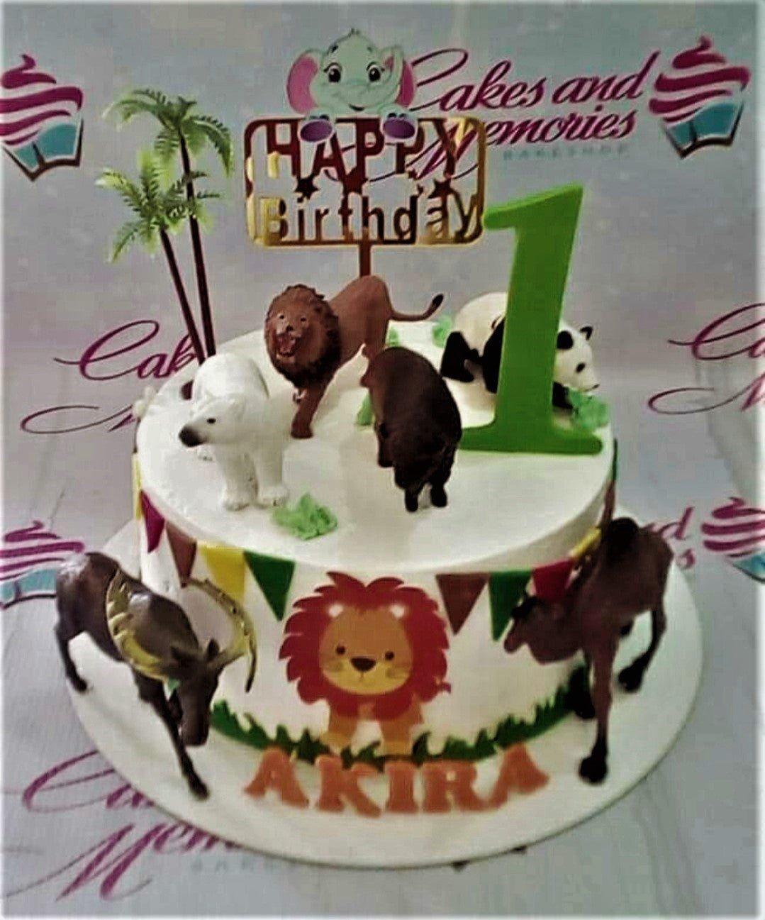 Safari themed birthday cake with plastic animal toys, palm trees, and a gold Happy Birthday topper for Akira's 1st birthday.