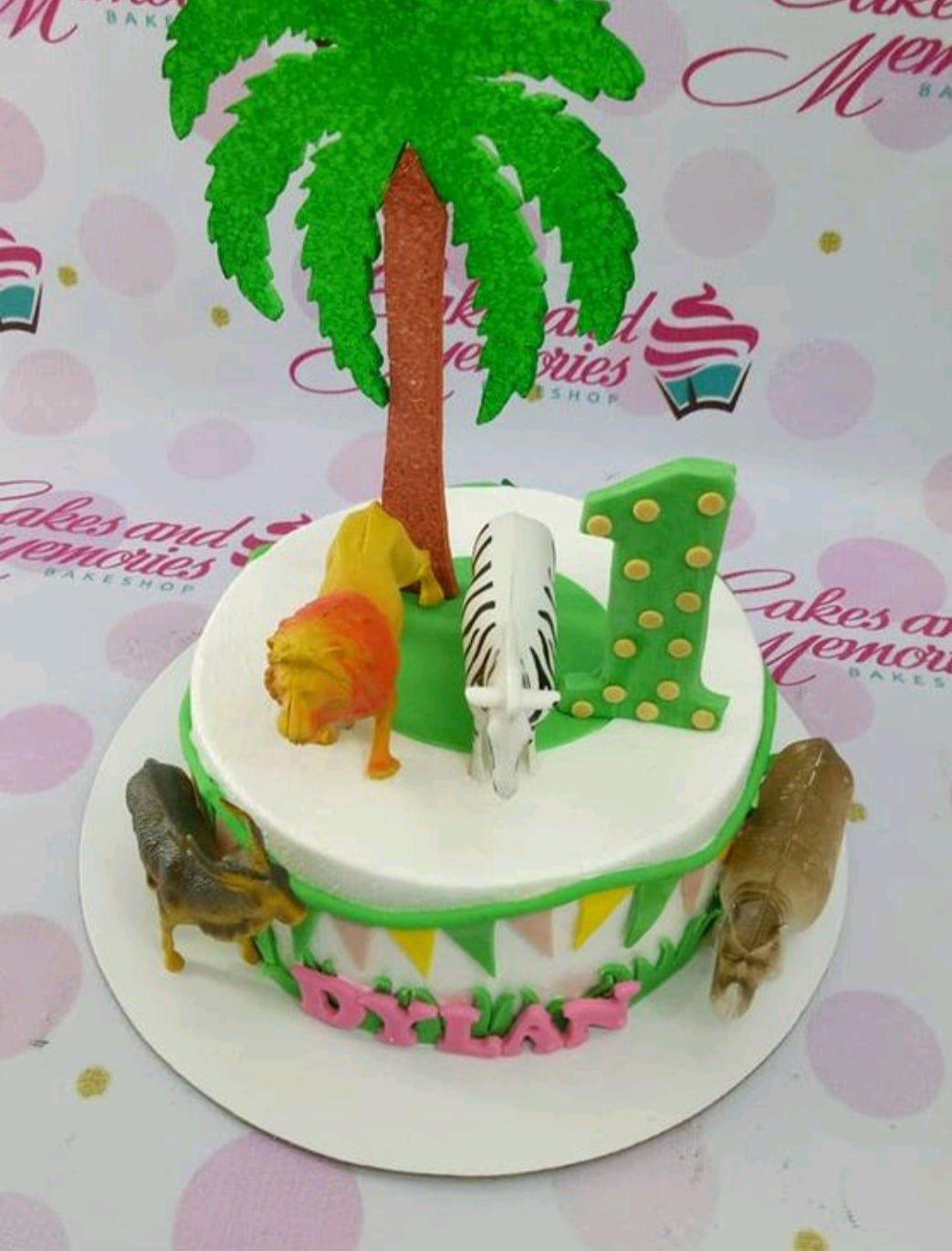Safari themed birthday cake with a palm tree, lion, zebra, and hippo figures on white icing with green grass accents.