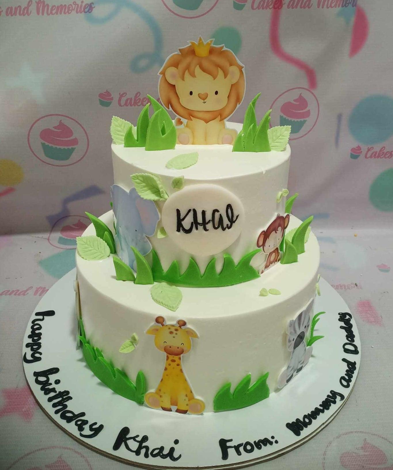 Two-tier white safari animal cake with a lion king topper, giraffe, zebra, elephant, and monkey printouts, and green fondant grass and leaves.