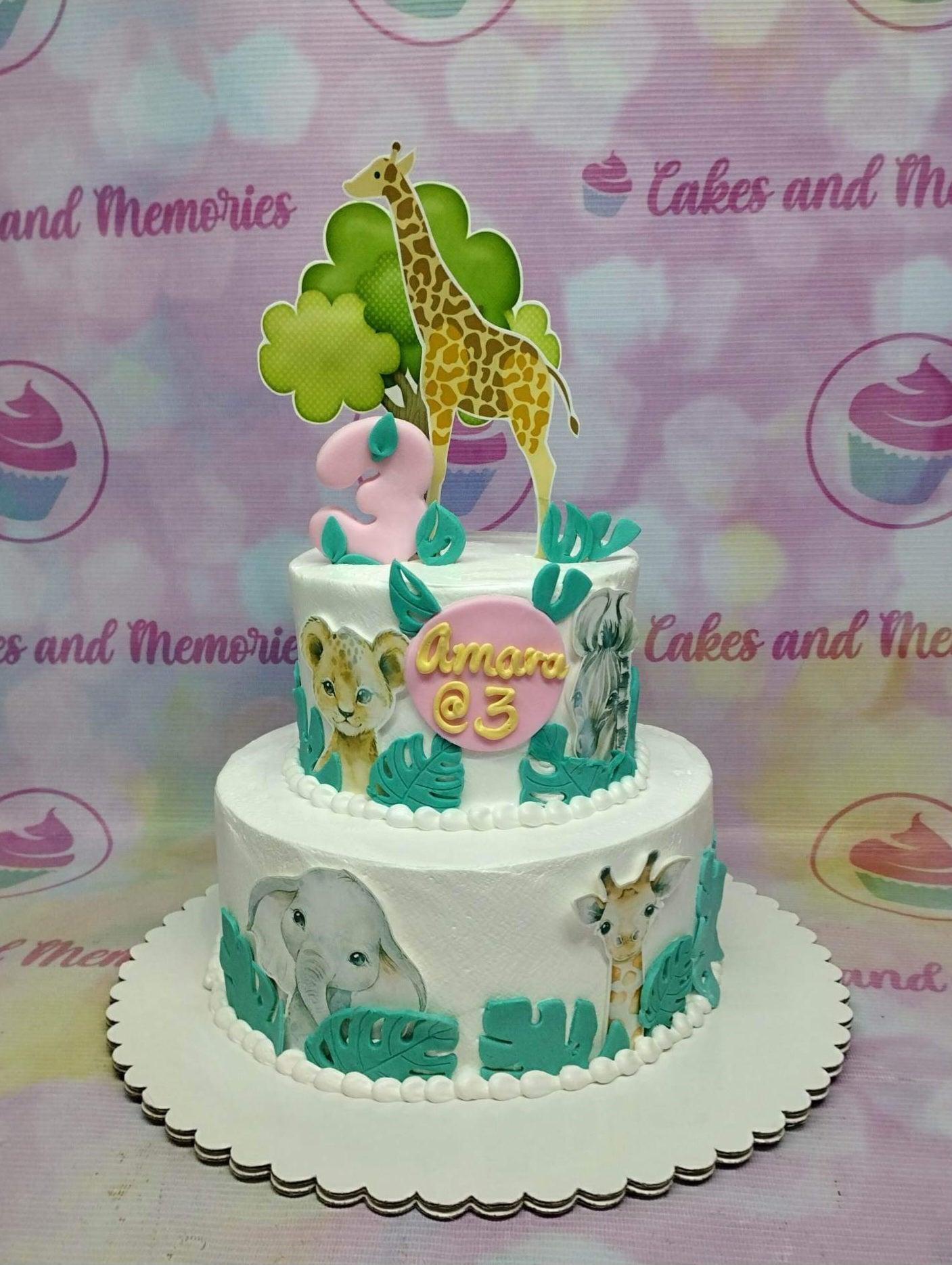 Two-tier white safari animal cake with giraffe, elephant, lion, and zebra printouts, green leaves, and a pink number 3 topper.