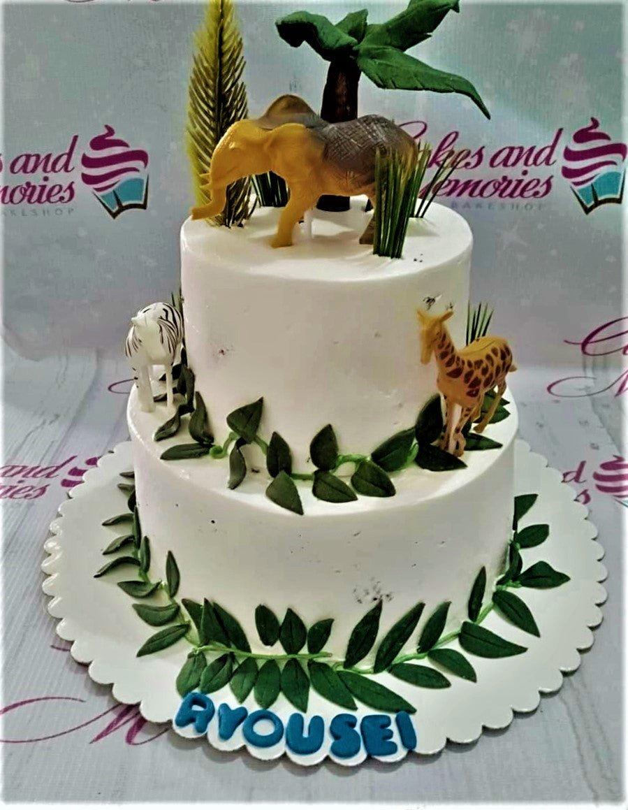 Two-tier white safari themed cake with plastic toy animals including an elephant, giraffe, and zebra, decorated with green fondant leaves.
