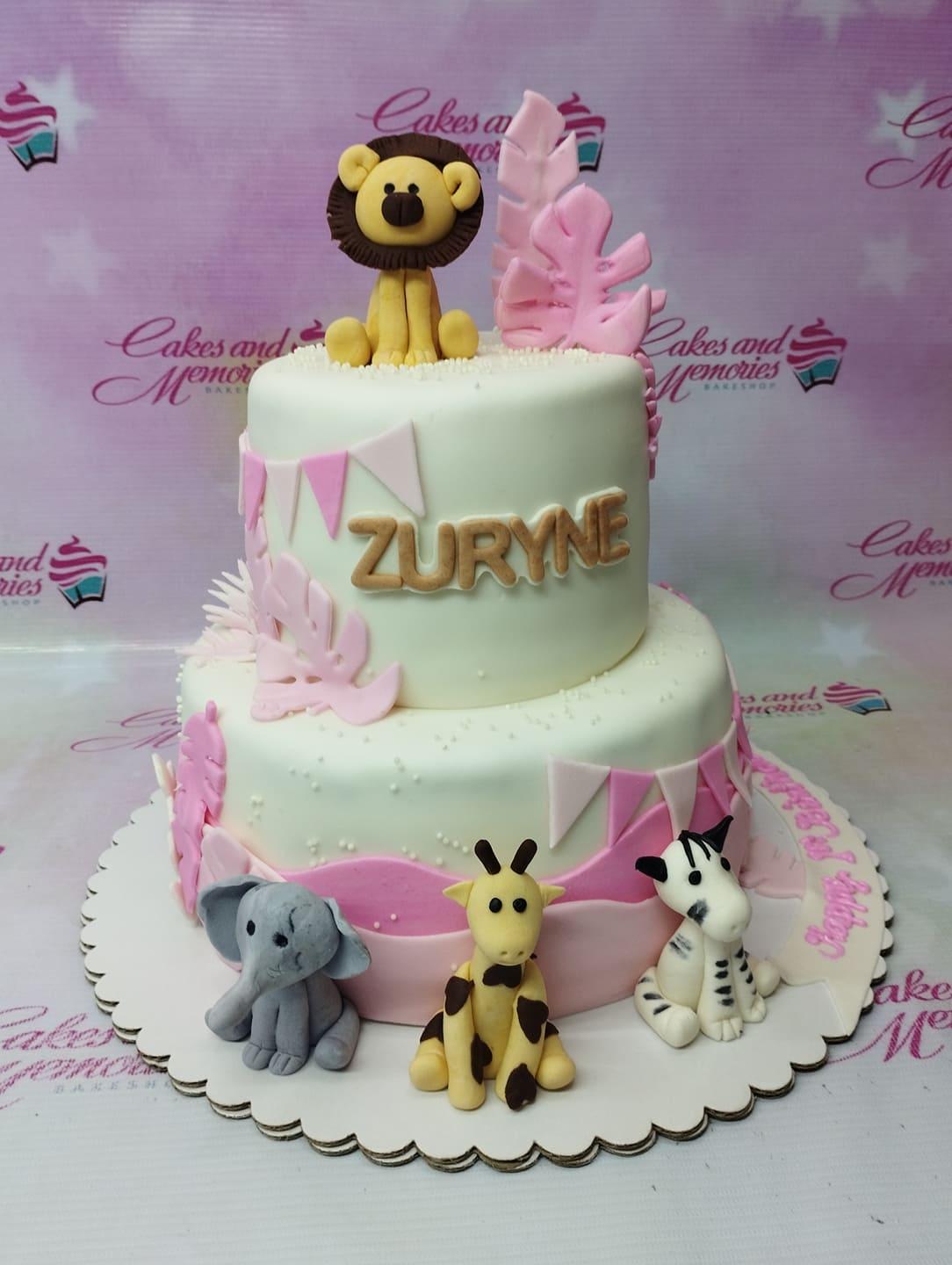 Two-tier white fondant safari cake with pink accents, featuring a fondant lion, giraffe, elephant, and zebra with pink tropical leaves.