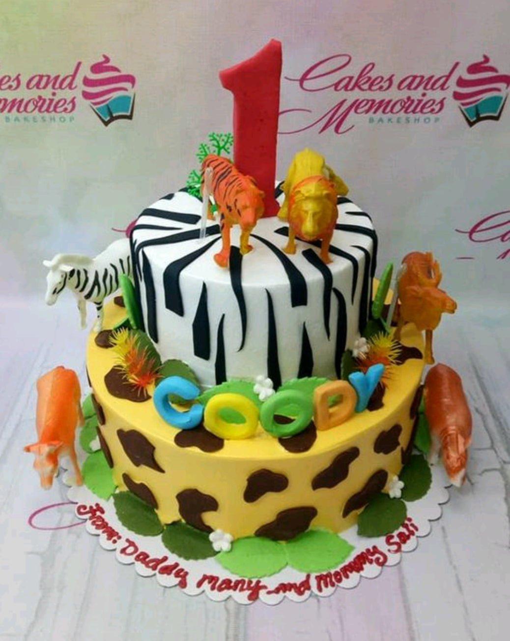Two-tier safari animal birthday cake with zebra and giraffe patterns, featuring toy animals and a large red number one topper.