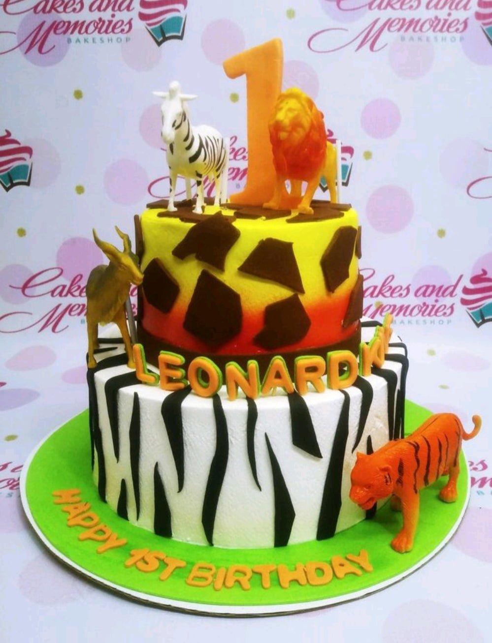 Two-tier safari themed birthday cake with zebra stripes, giraffe patterns, and plastic animal figurines including a lion, tiger, and zebra.