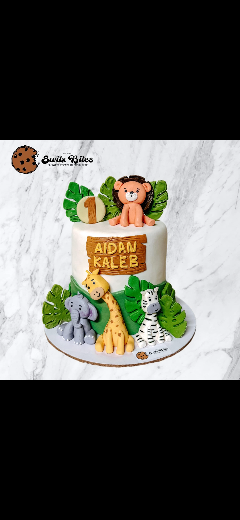 Safari themed 1-tier fondant cake with 3D edible animals including a lion, giraffe, elephant, and zebra with tropical leaves and a wooden name plaque.