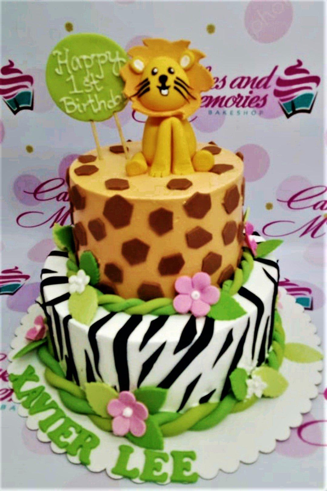 Two-tier safari themed birthday cake with a fondant lion topper, giraffe and zebra patterns, and pink flowers.