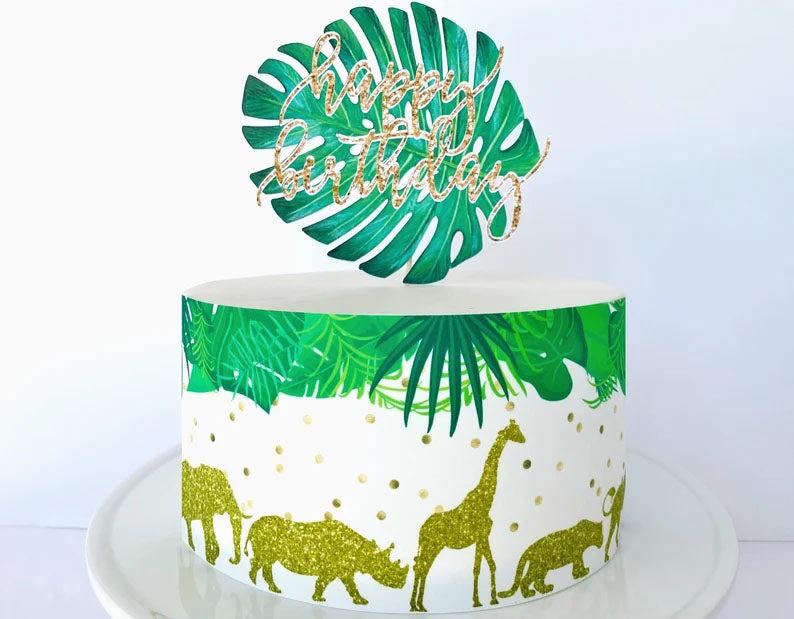 Safari themed birthday cake with tropical leaf topper and gold glitter animal silhouettes on a white icing base.