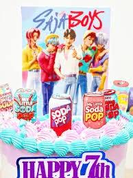 Pink K-pop themed cake with Saja Boys character printouts, soda can toppers, and blue and pink icing rosettes.