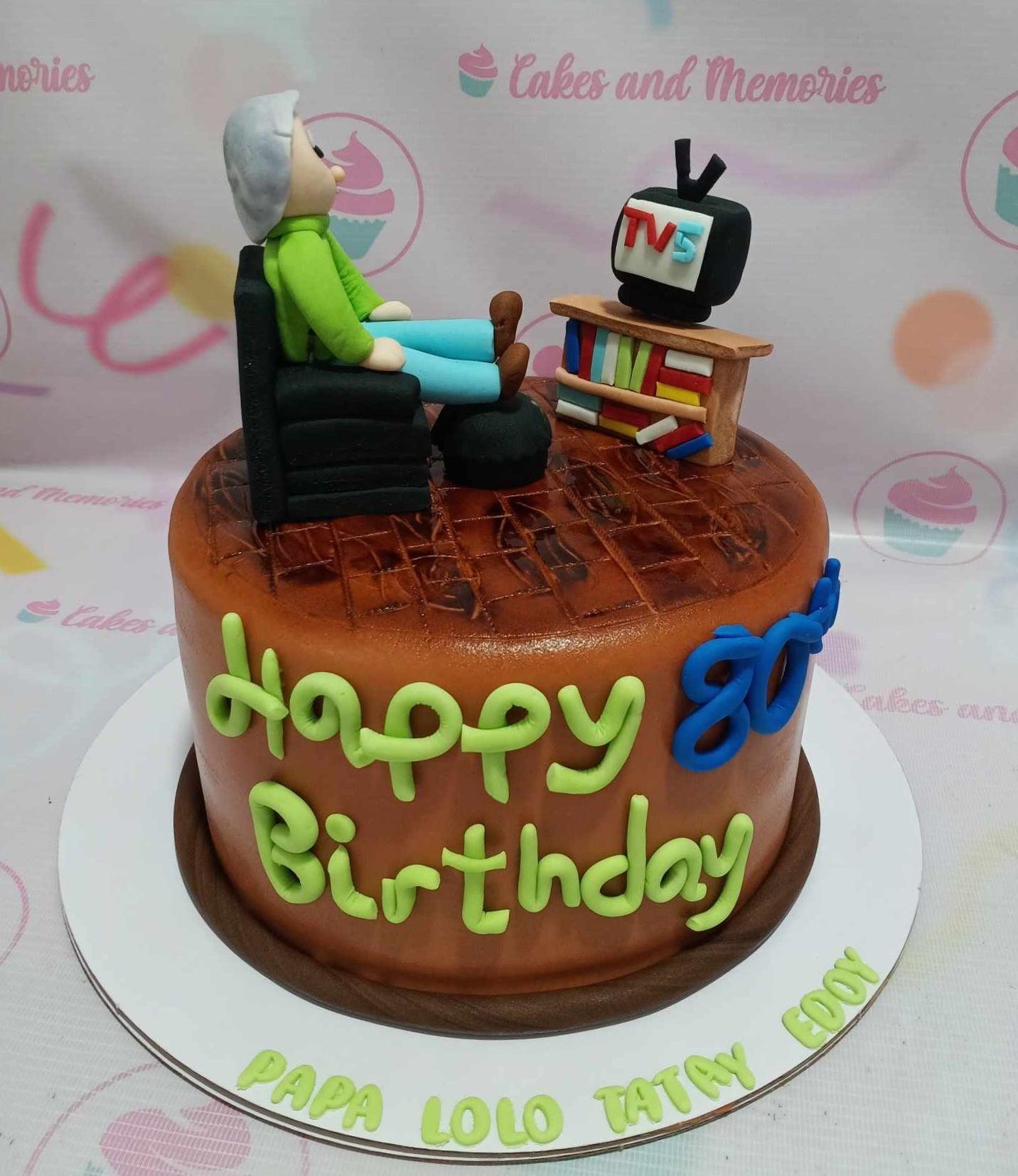 Brown fondant cake for an 80th birthday featuring a 3D fondant man in a chair watching a TV on a bookshelf topper.
