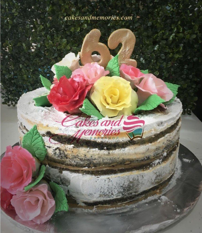 Naked chocolate cake with white icing, topped with large gold number 62 and a bouquet of pink, red, and yellow edible roses.