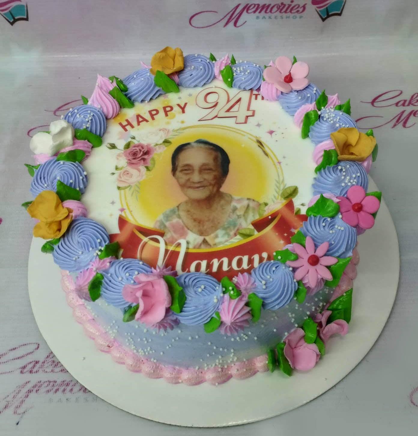Round birthday cake with a photo print of an elderly woman, purple and pink icing rosettes, and yellow edible flowers.