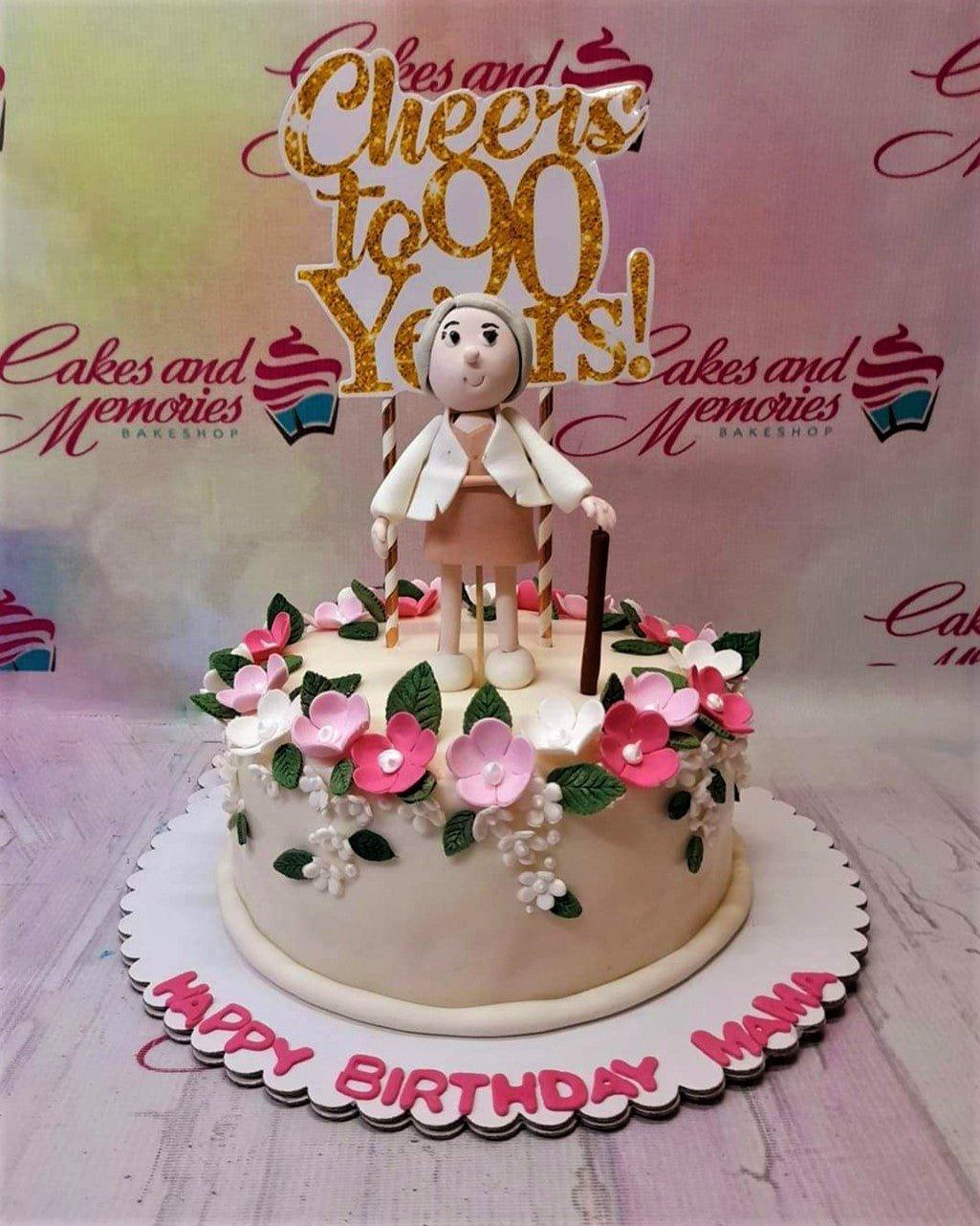 White fondant birthday cake for a 90th birthday featuring a 3D elderly woman figure, pink and white flowers, and a gold glitter topper.