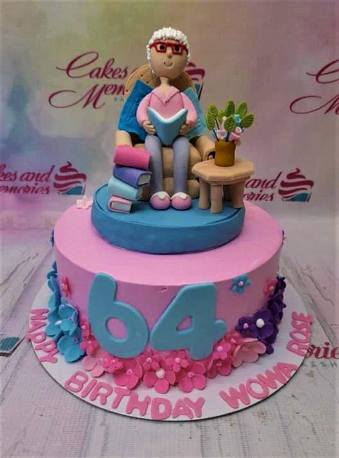 Pink fondant birthday cake for a 64th birthday featuring a detailed edible 3D figure of a grandmother reading a book in a chair.
