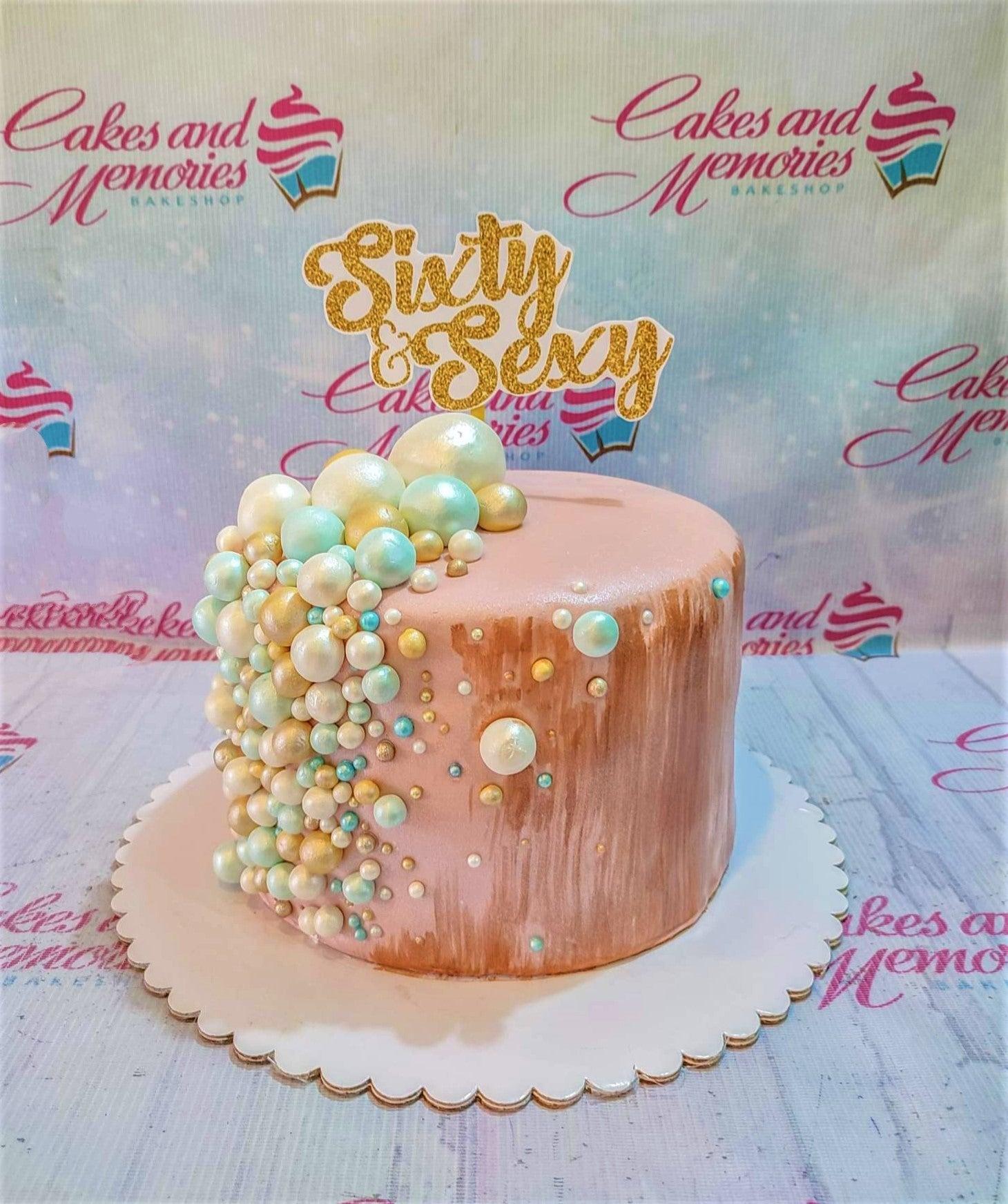 Elegant pink fondant cake with a gold glitter Sixty and Sexy topper and a cascade of white, gold, and teal edible pearls.