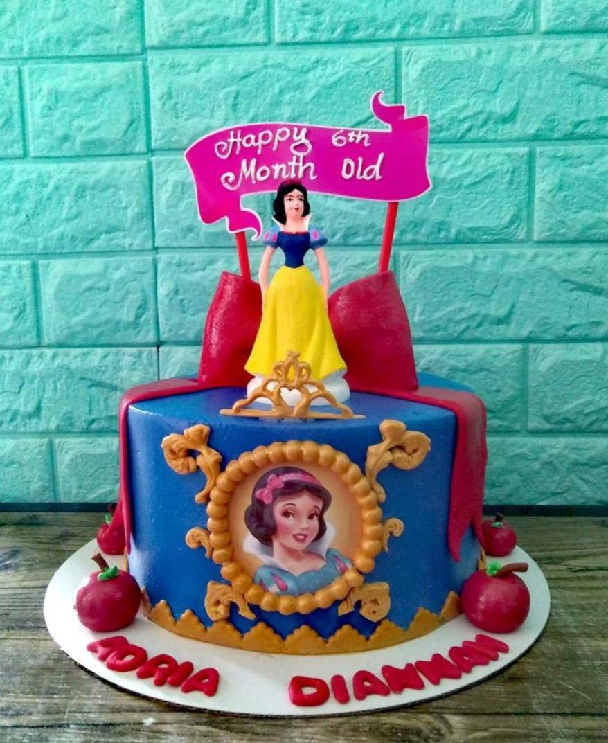 Blue Snow White themed cake with a Snow White figurine, red apples, and a pink Happy 6th Month Old banner.