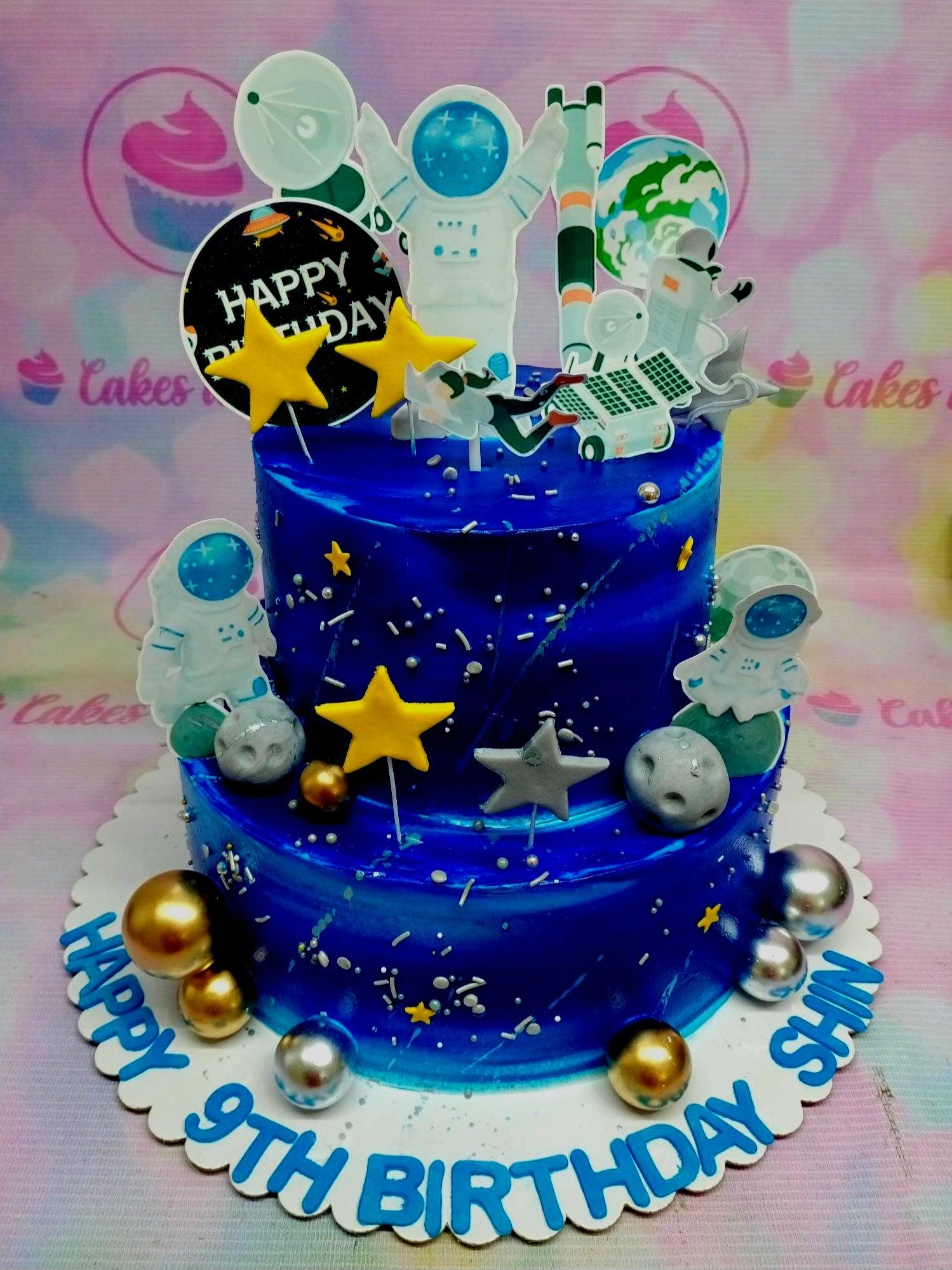 Two-tier deep blue space-themed birthday cake with astronaut and planet printouts, yellow stars, and silver dragees.