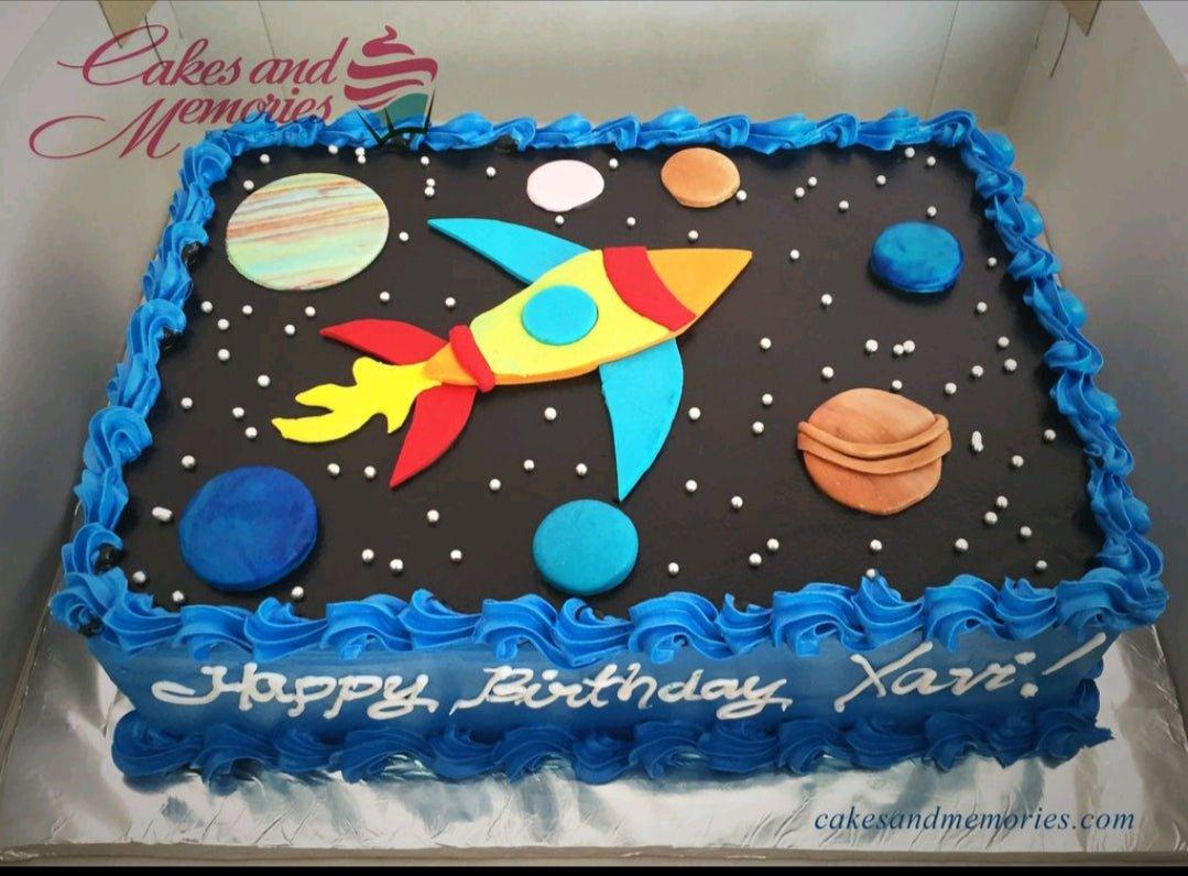 Space themed rectangle cake with a rocket ship, planets, and silver dragees on black icing with blue borders.
