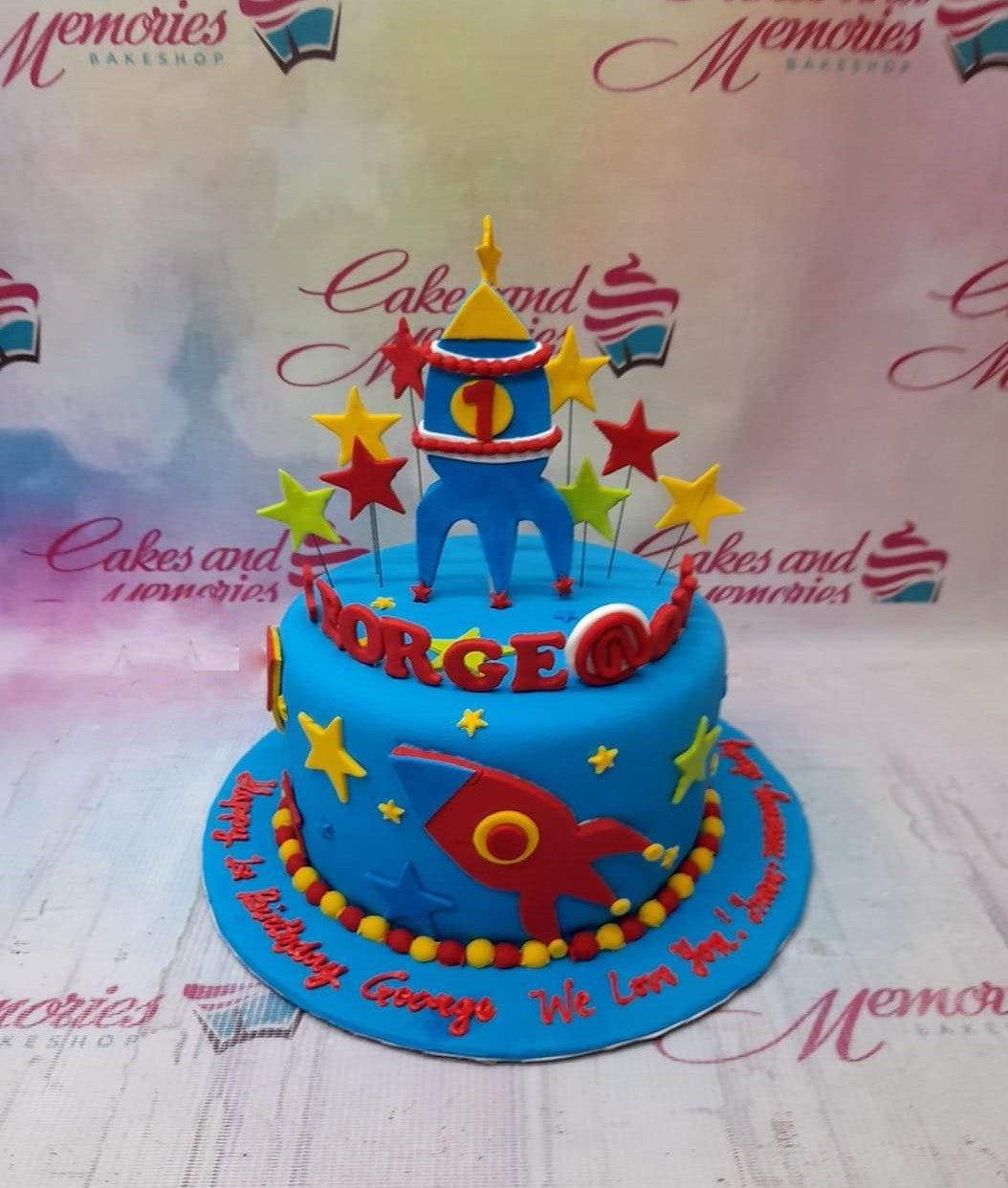 Blue space-themed rocket ship cake with yellow stars and red accents for a first birthday celebration.