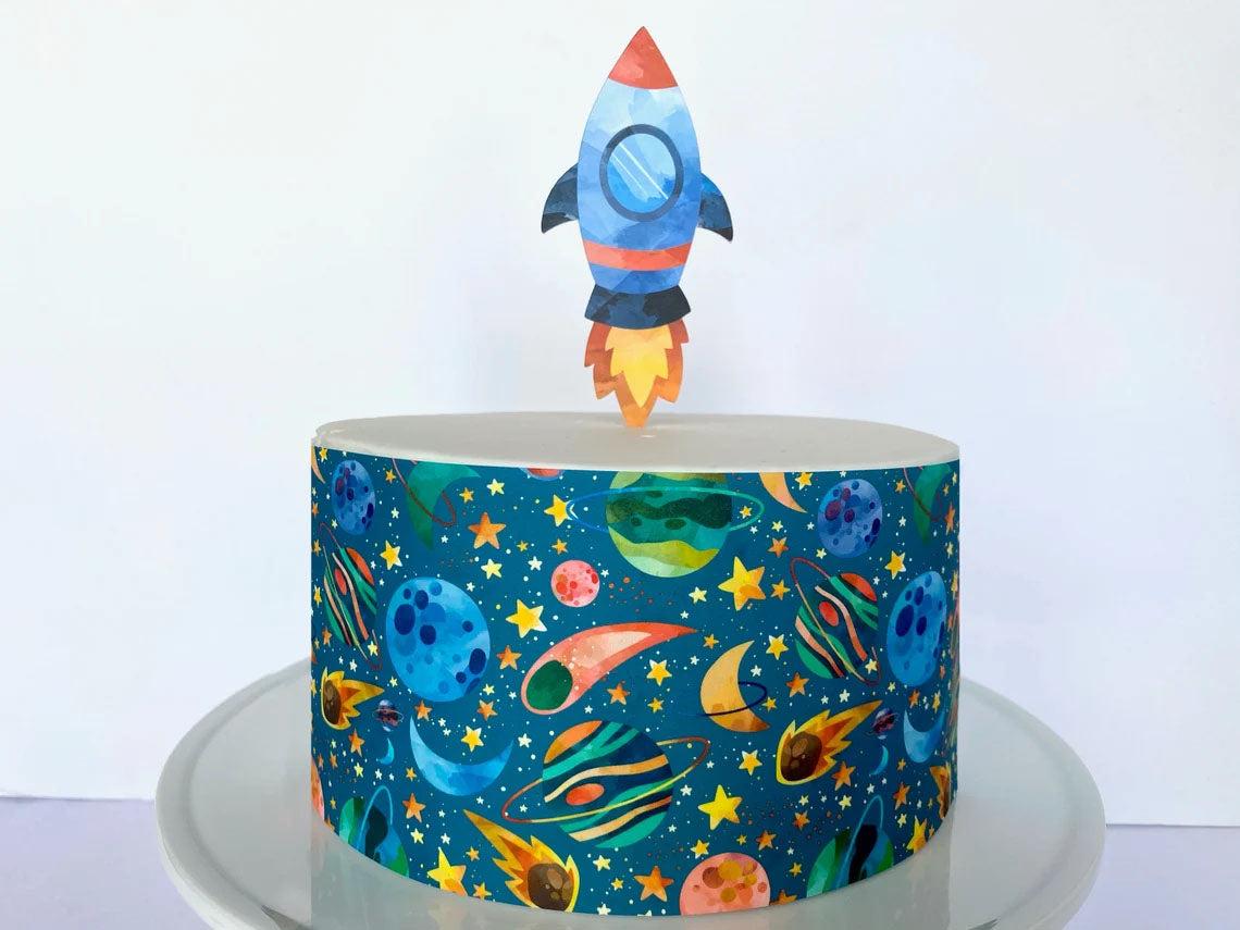 Space themed birthday cake with a rocket ship topper and a colorful galaxy planet side wrap on white icing.