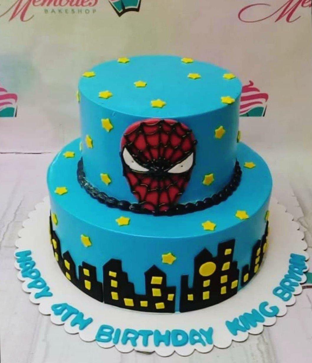 Blue 2-tier Spiderman birthday cake with a fondant Spiderman mask, city skyline silhouette, and yellow stars.