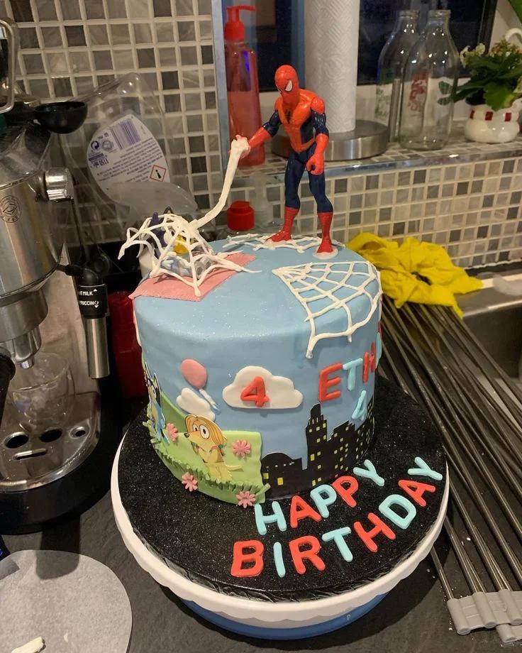 Blue Spiderman and Bluey themed birthday cake with a Spiderman toy figure and fondant city skyline.