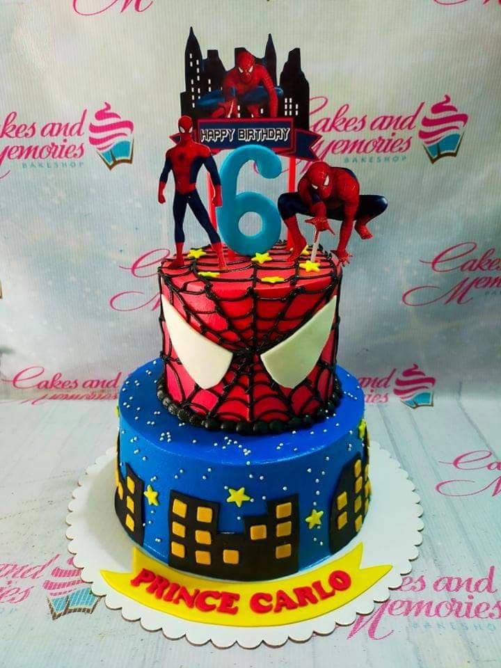 Two-tier Spiderman themed birthday cake with blue city skyline bottom tier and red webbed top tier featuring Spiderman character cutouts.