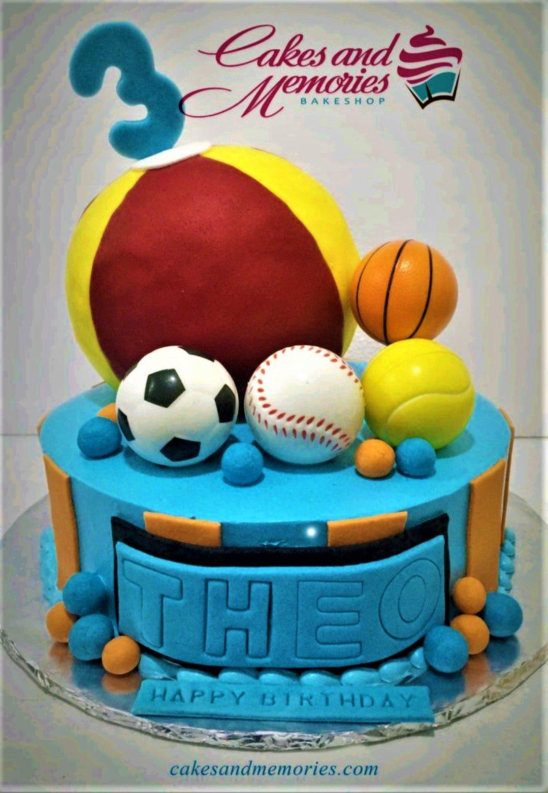 Blue sports themed birthday cake for Theo featuring a large beach ball, soccer ball, baseball, basketball, and tennis ball toppers.