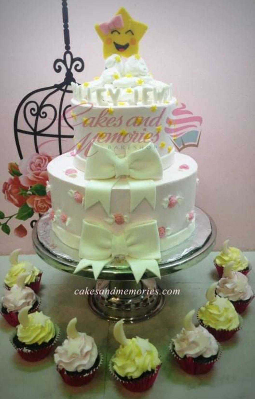 Two-tier white cake with yellow star topper, pink bows, and small pink rosettes, surrounded by matching cupcakes.
