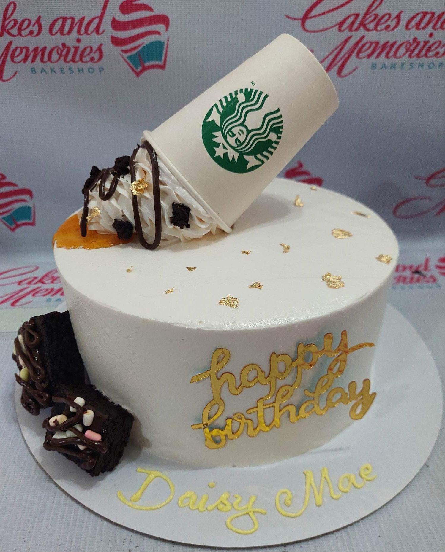 White Starbucks themed cake with a tilted coffee cup topper, chocolate drizzle, brownie pieces, and gold leaf accents.