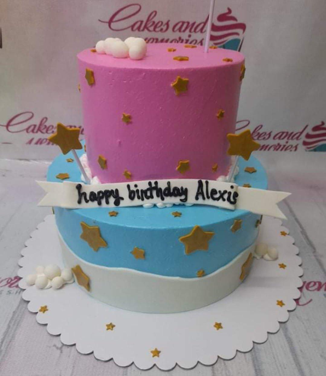 Two-tier birthday cake with pink top tier and blue bottom tier, decorated with gold stars and white fondant clouds.