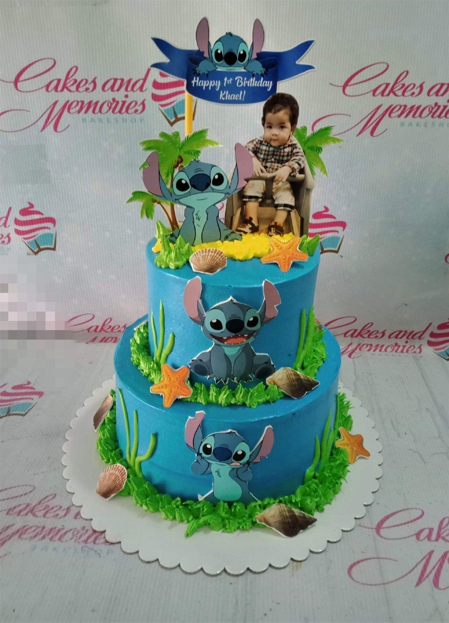 Two-tier blue Stitch themed birthday cake with character printouts, green icing grass, and sea shell decorations.