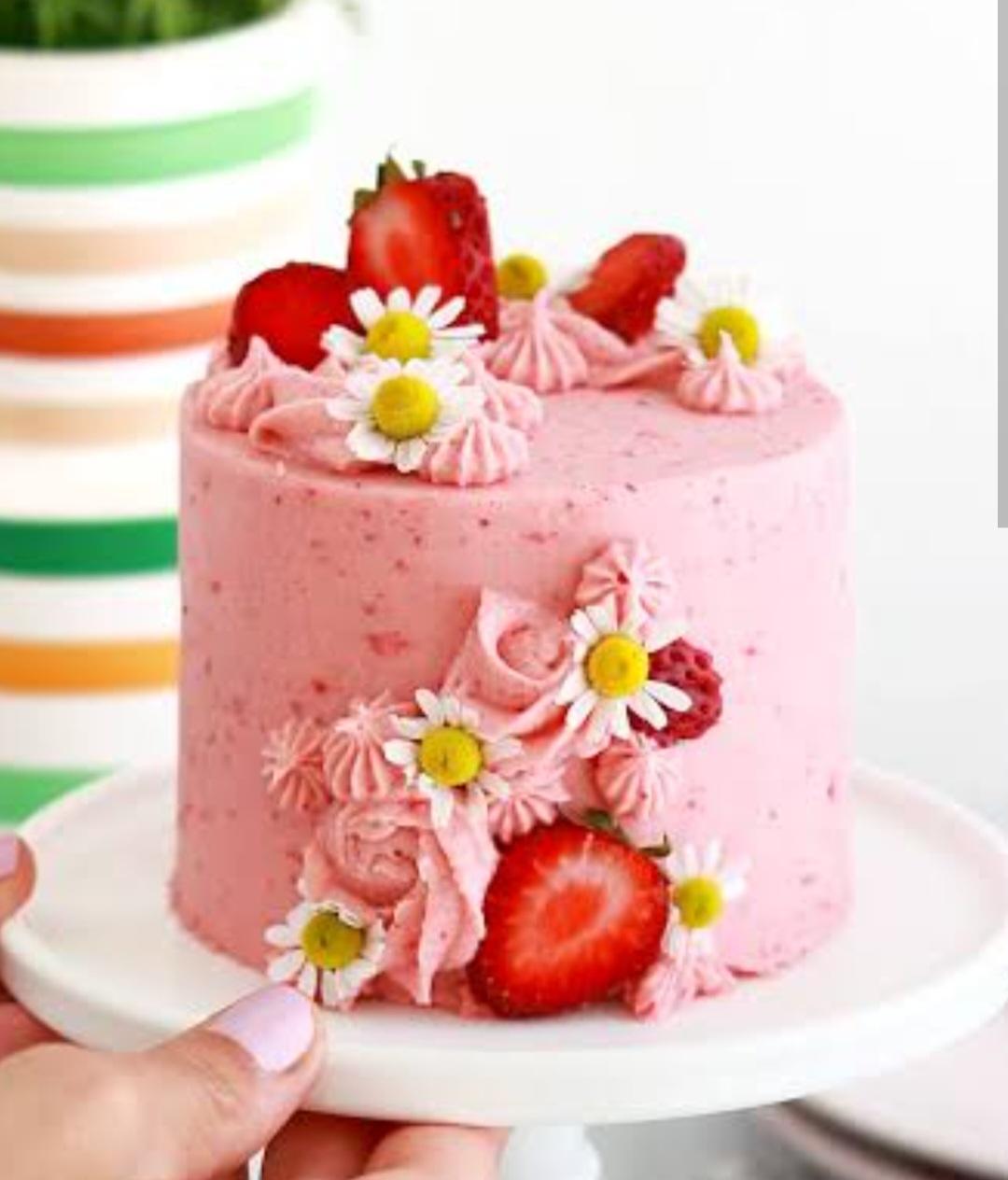 Small pink strawberry cake with fresh strawberry slices, white daisies, and pink icing rosettes on a white cake stand.