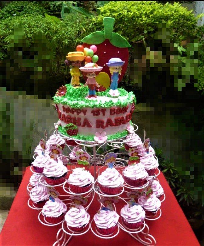 Strawberry Shortcake themed birthday cake with 3D character figures, a large strawberry backdrop, and pink cupcakes on a white stand.