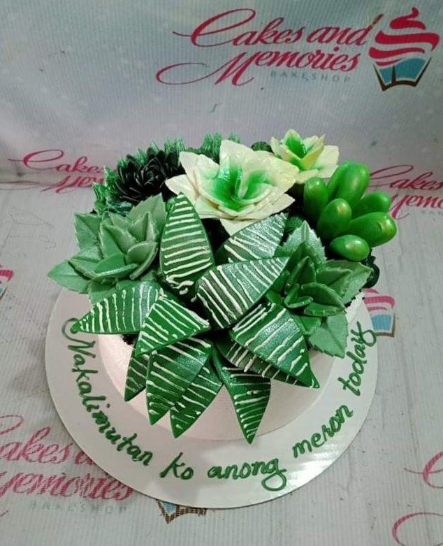 A round white cake topped with a dense arrangement of green and white succulent-shaped icing decorations and a message on the base board.