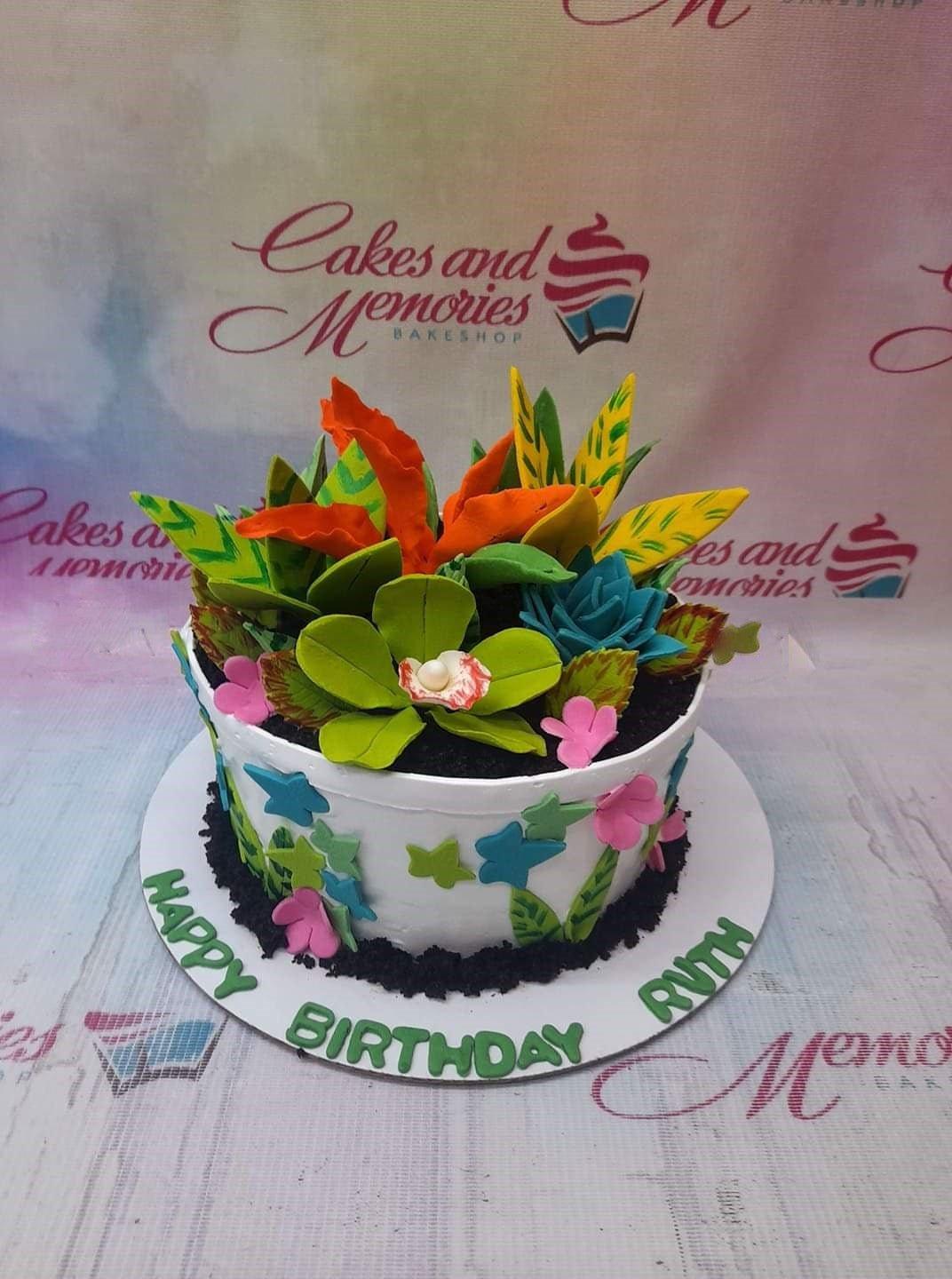A white round cake decorated with vibrant green, orange, and blue succulent-style fondant toppers and pink flowers on a black crumb base.
