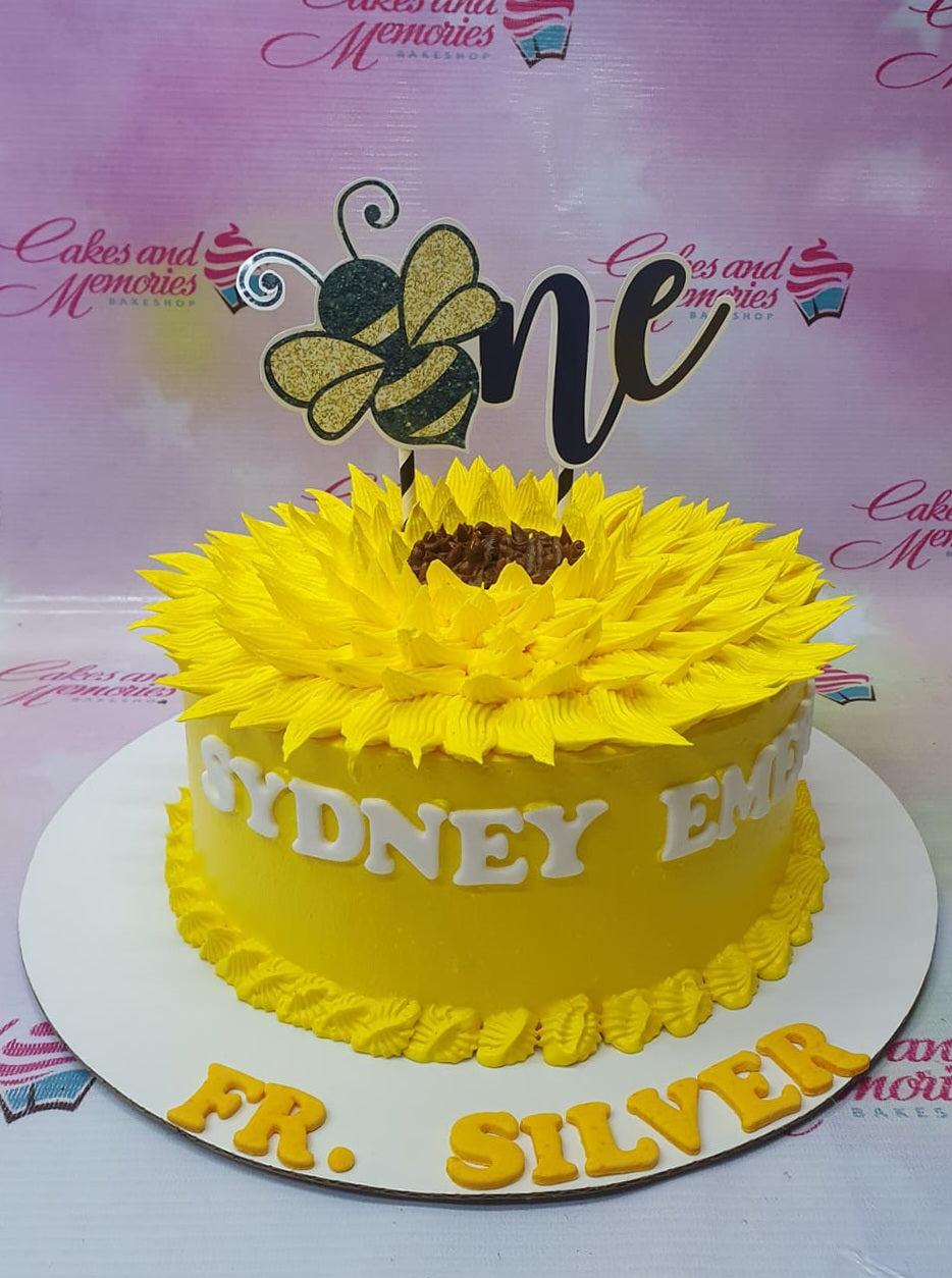 Yellow sunflower themed birthday cake with a glittery bee and 'one' topper, featuring yellow icing petals and white gumpaste name letters.