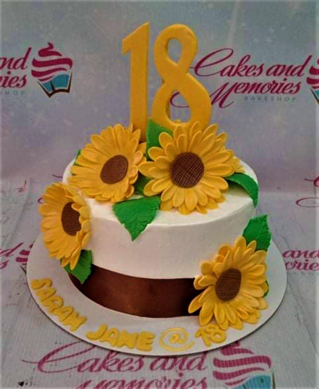 White and brown 18th birthday cake decorated with large yellow gumpaste sunflowers and green leaves with a large yellow number 18 topper.