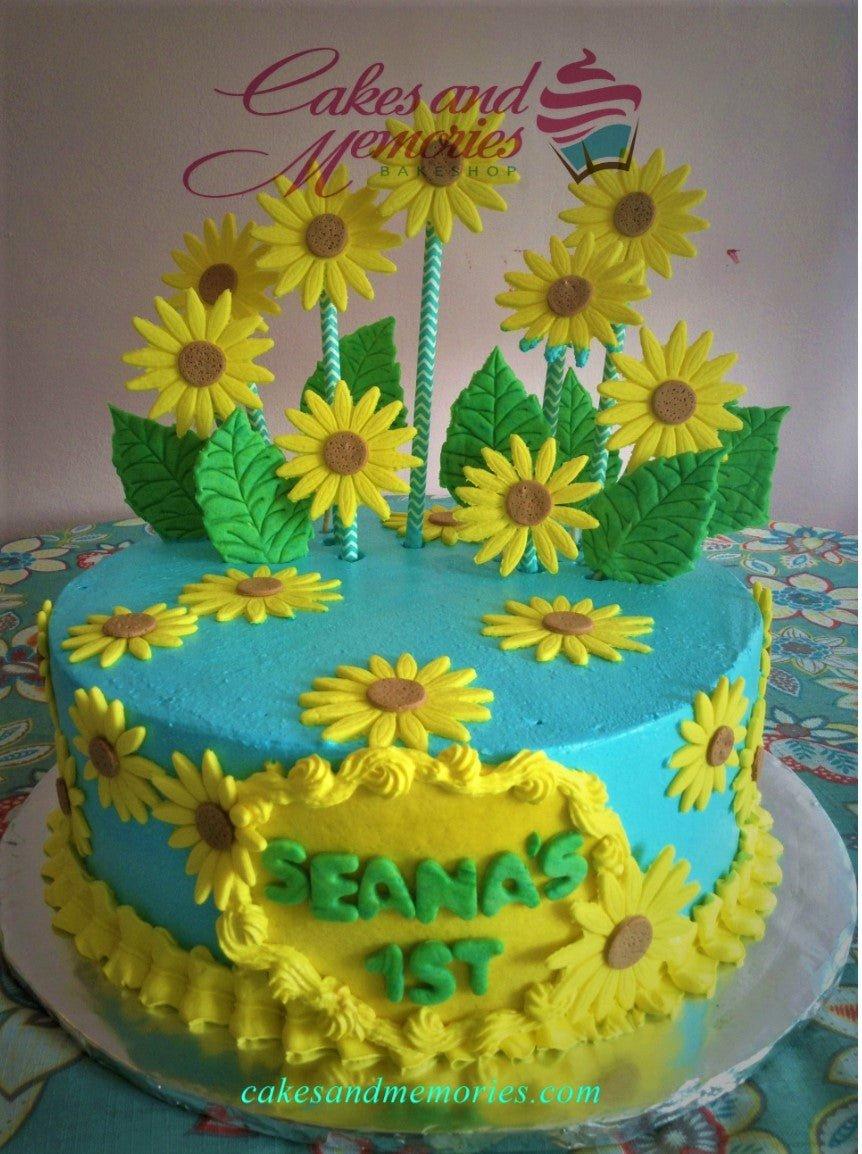 Light blue sunflower themed birthday cake with yellow fondant sunflowers and green leaves on a yellow icing plaque.