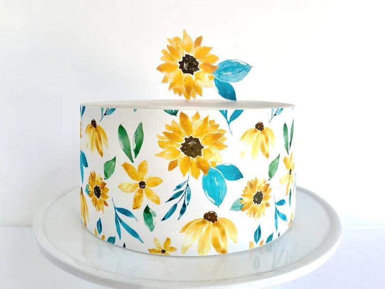 White round cake with watercolor sunflower and teal leaf patterns on the sides and a sunflower topper.