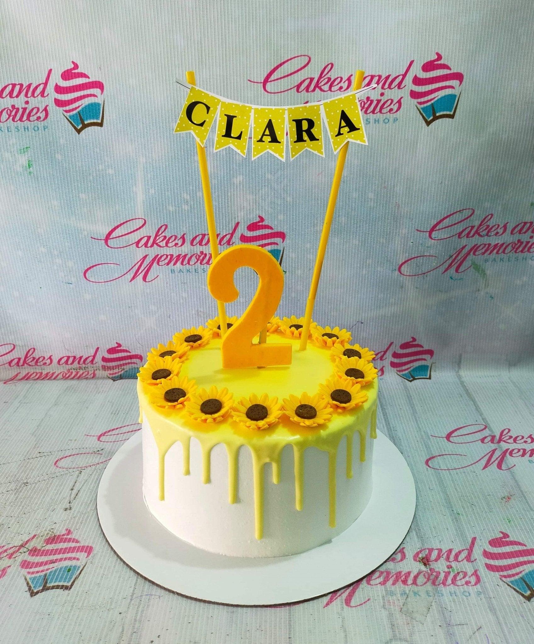 White 1-tier sunflower birthday cake with yellow drip and a custom name banner for Clara's 2nd birthday.