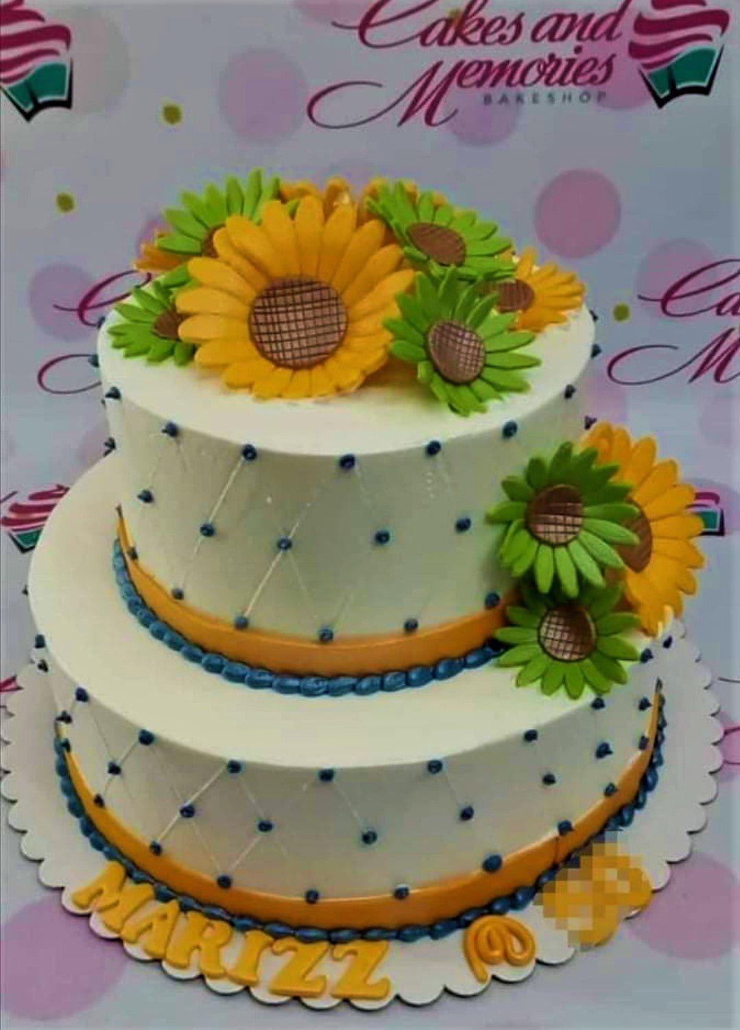 Two-tier white cake decorated with large yellow and green sunflowers, blue icing dots, and orange ribbon borders.