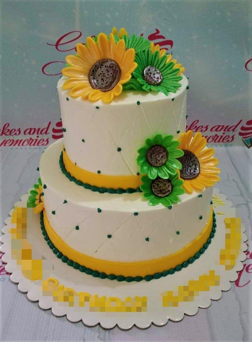 Two-tier white cake decorated with large yellow and green fondant sunflowers and dark green icing dots.