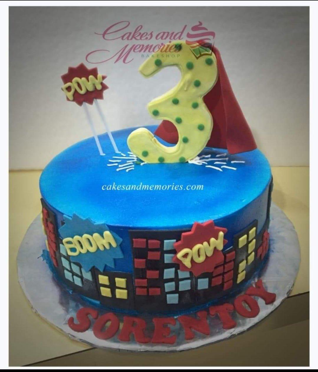 Blue superhero themed birthday cake with a large yellow number 3 topper, red cape, and city skyline fondant decorations.