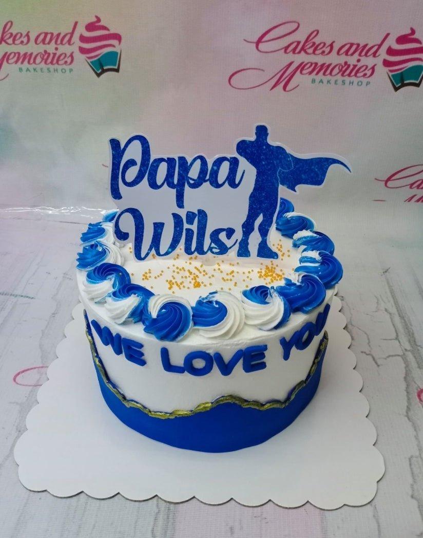 White 1-tier cake with blue and white icing swirls, gold sprinkles, and a blue superhero Papa Wils printout topper.