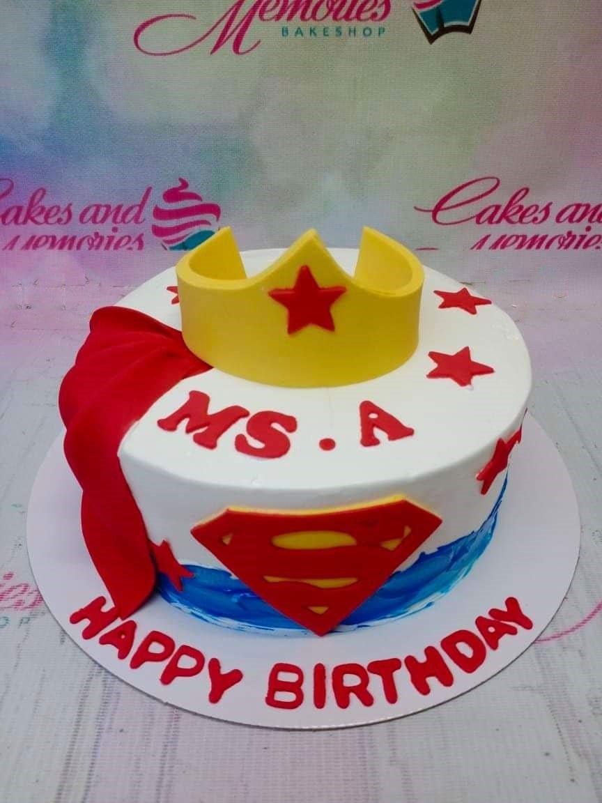 Superman and Wonder Woman themed birthday cake with a yellow fondant crown, red cape, and Superman logo on white icing.