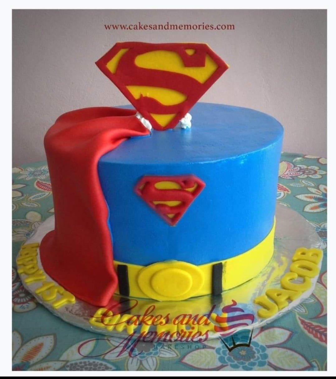 Superman themed blue fondant cake with a red cape, yellow belt, and Superman logo toppers for a 1st birthday.